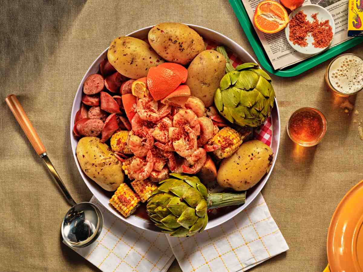 A round pan heaped with shrimp and artichokes, sliced sausage, orange slices, and corn. 