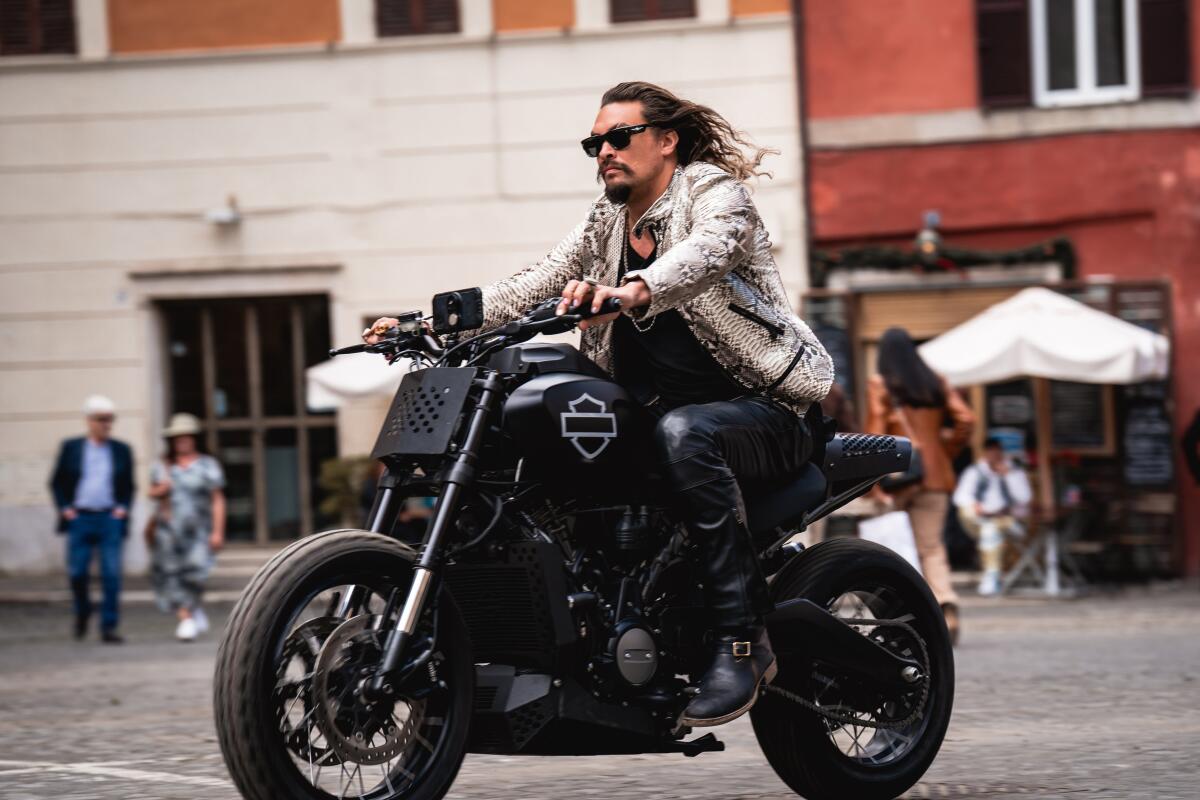 Jason Momoa rides a motorcycle down a cobblestone street.