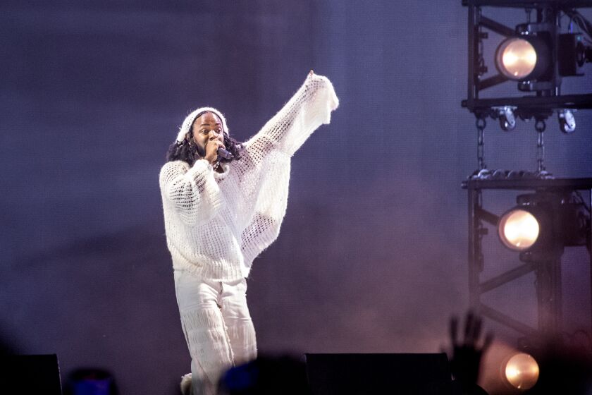 A man rapping into a microphone and raising his arm in an all-white outfit on a stage