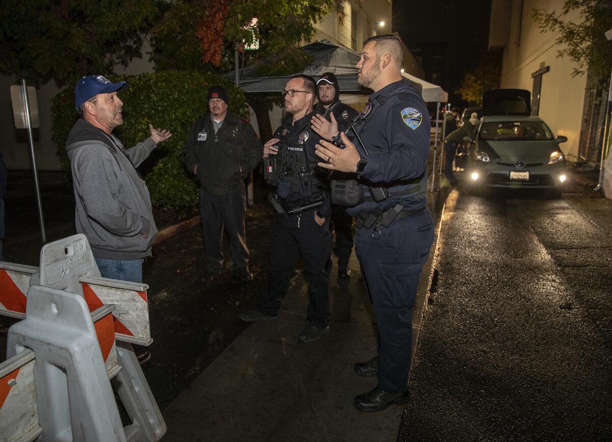 A man talks to people in uniforms in an alley at night.