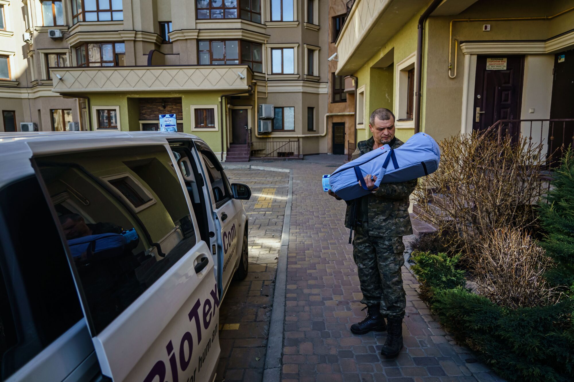 A man in army fatigues looks into a baby carrier he is holding while outside near a white van with its door open.