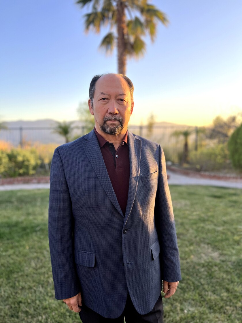 A man in a suit with a palm tree behind him stares into the camera.