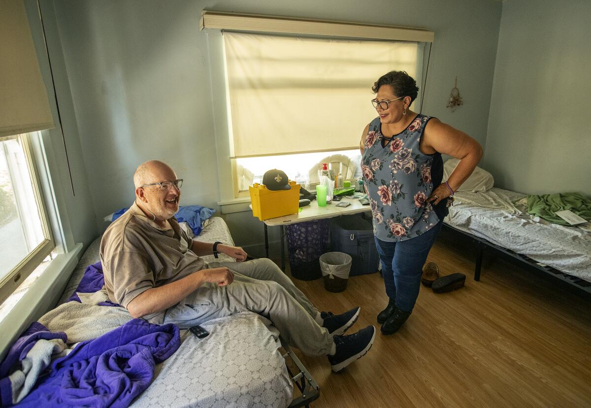 A woman standing in the middle of a room converses with a man seated on one of its two beds.