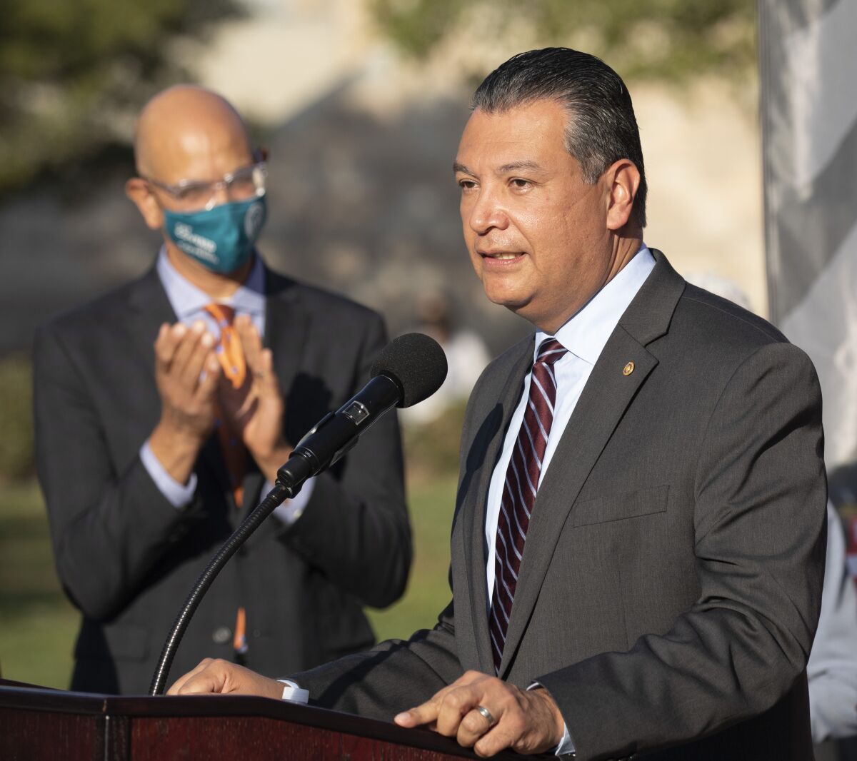 Alex Padilla speaks at a lectern while a man behind him applauds.