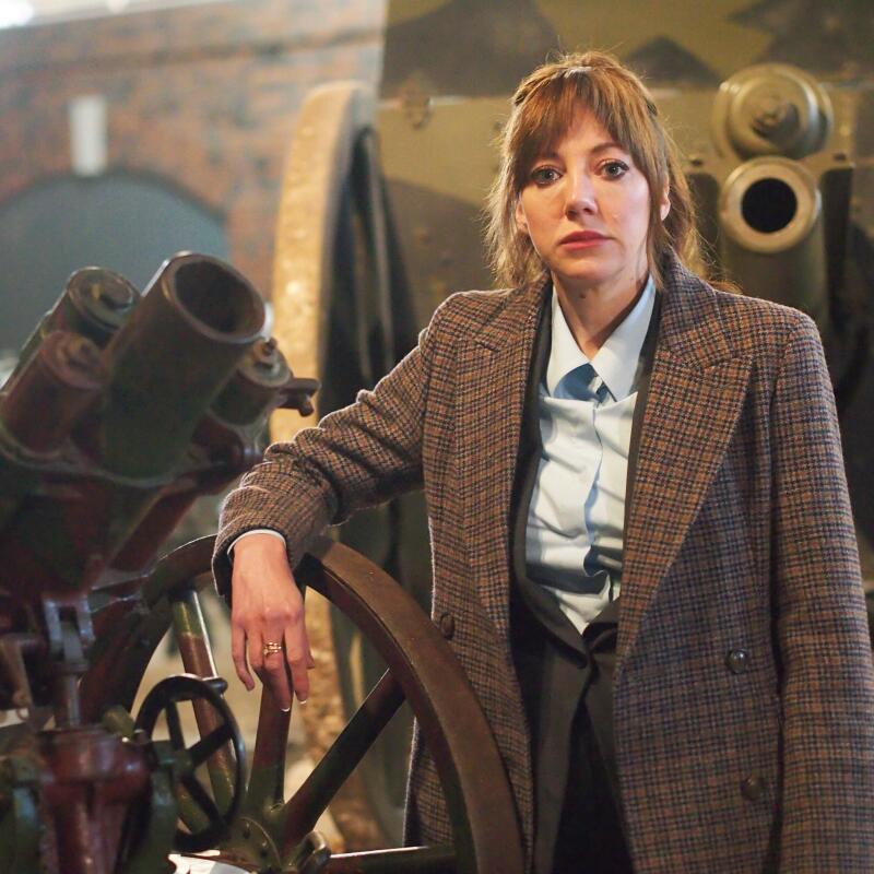 A woman in a tweed jacket leaning on an old cannon looking straight into camera