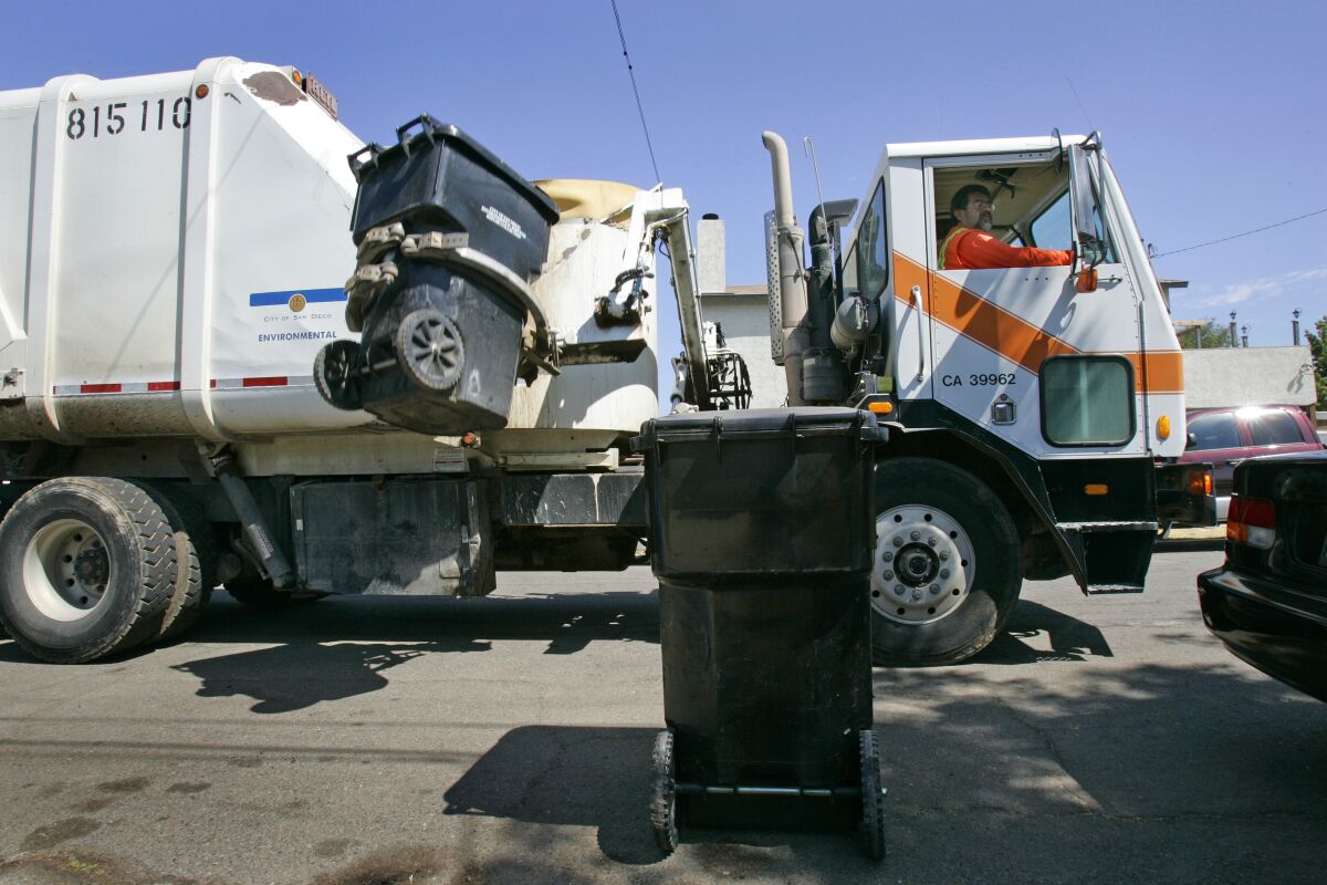 A garbage truck driver watches as his truck's hydraulic arm lifts a trash can to dump it into the truck.