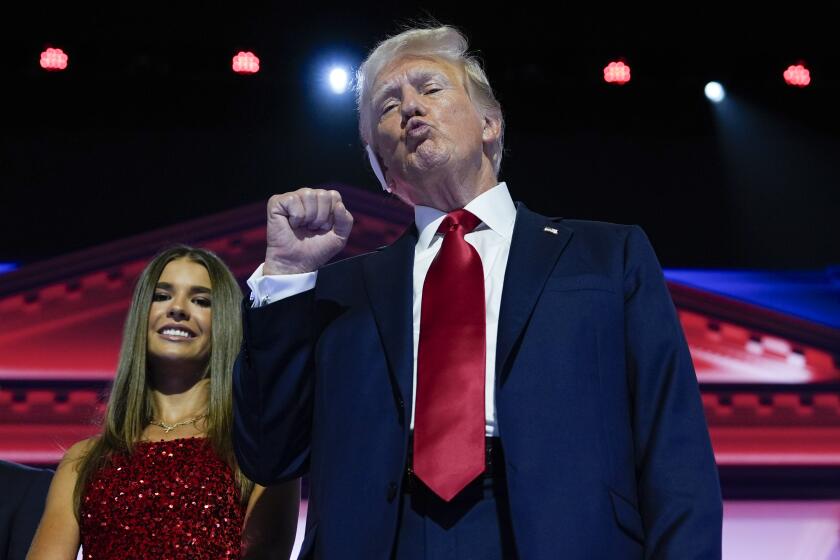 Trump in a suit and red tie while holding up a fist and pursing his lips. A young woman in a red sparkly dress is behind him.