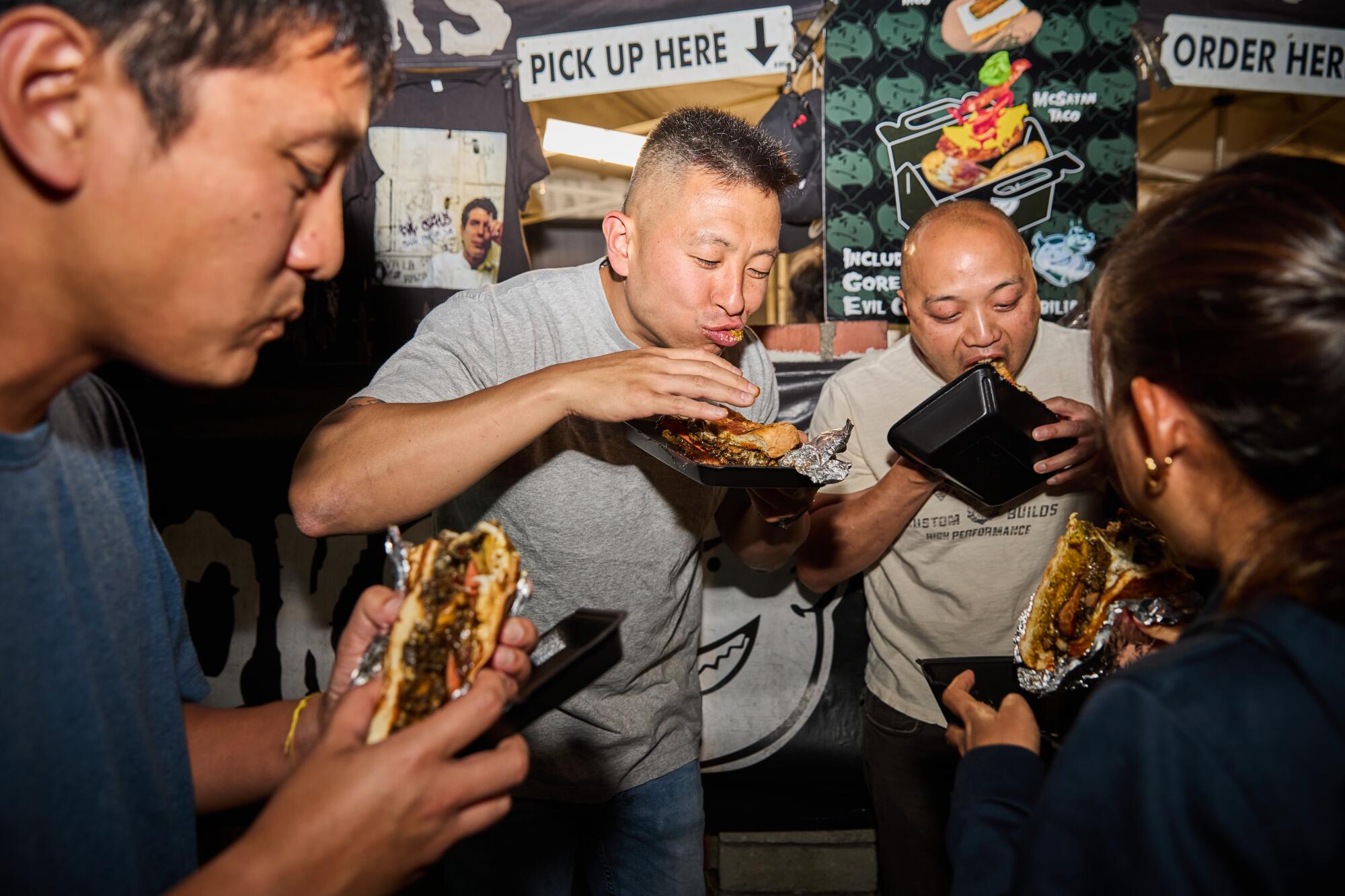 A group of diners stands eating tacos on the sidewalk.