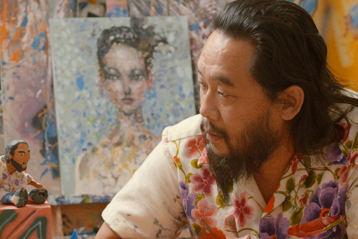  A bearded man in a colorful floral shirt sits in front of a painting of a woman.
