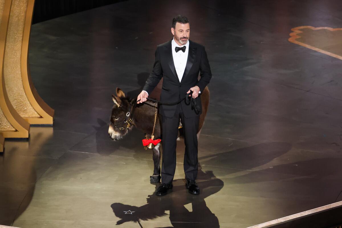 A man wearing formalwear holds the leash on a donkey onstage.