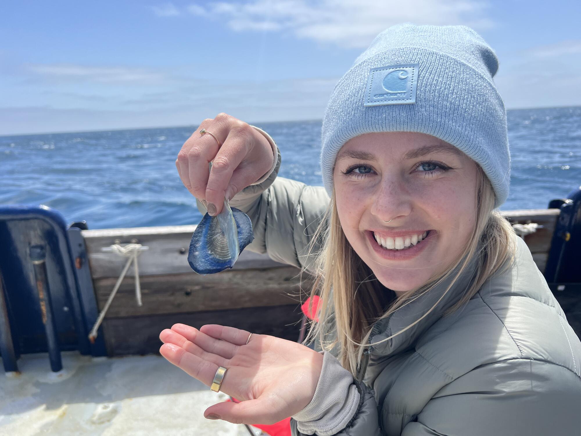 A woman on a boat wearing a blue beanie and holding a Velella velella specimen