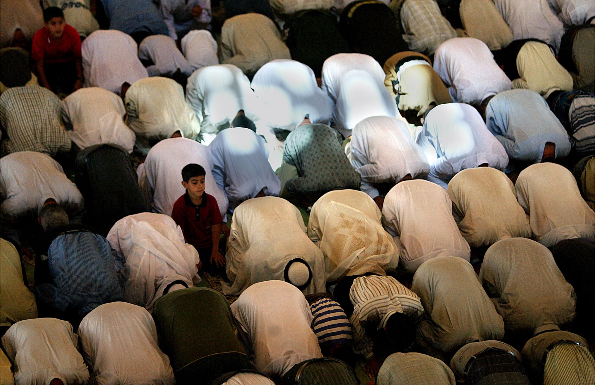 A boy sits up amid rows of people bowed over on the floor in prayer