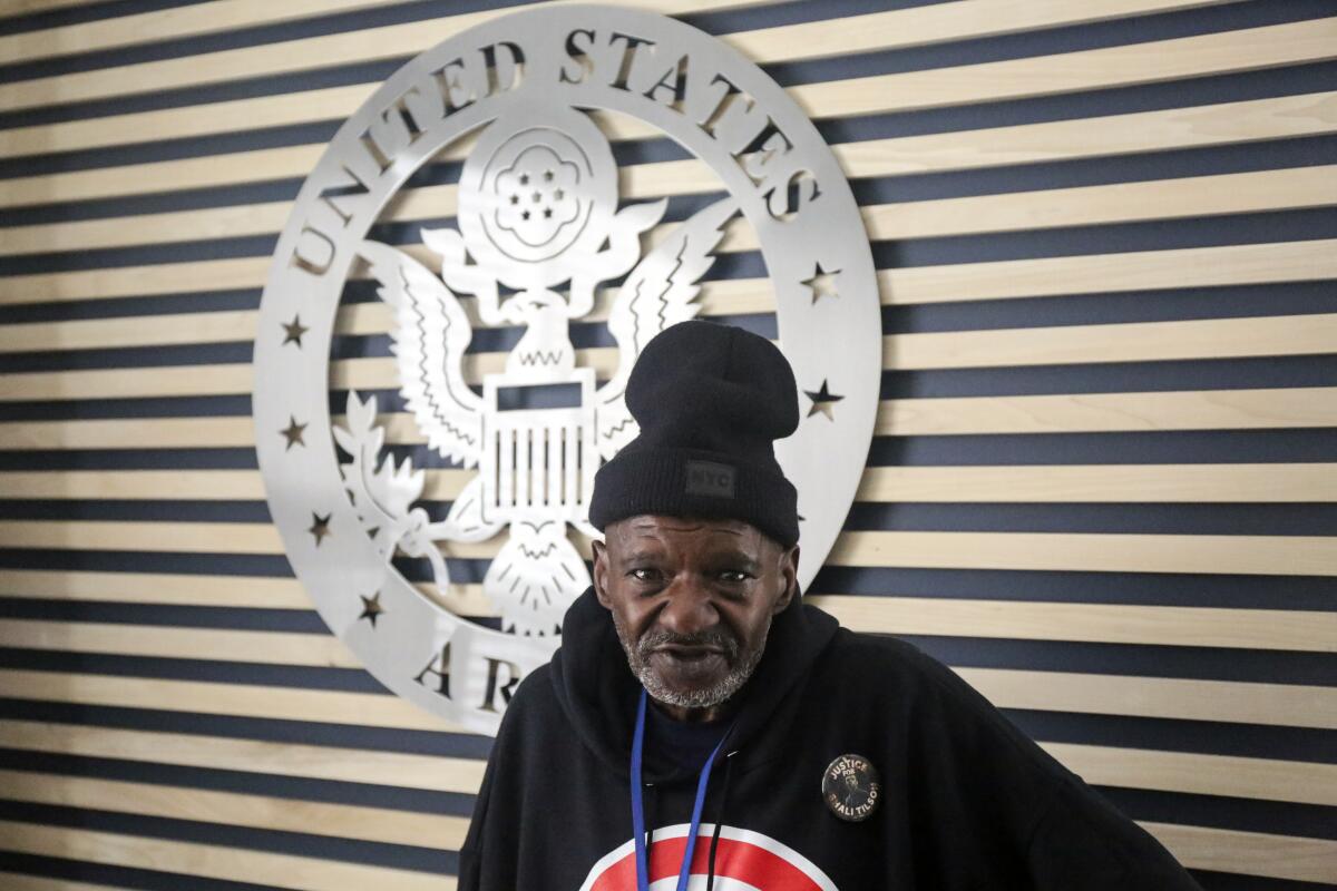 A man poses in front of a U.S. Army seal while wearing a sweatshirt and hat.