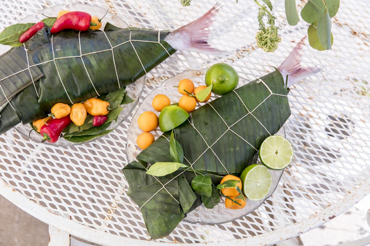 Only the tails can be seen of two fish wrapped in banana leaves, one on a plate with citrus; the other, with peppers.
