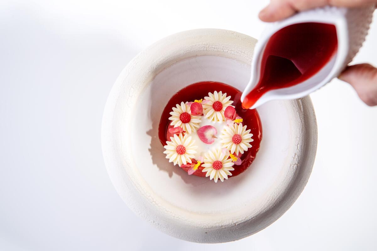 A hand pours red liquid into a bowl featuring white-and-pink flower-like dessert shapes.