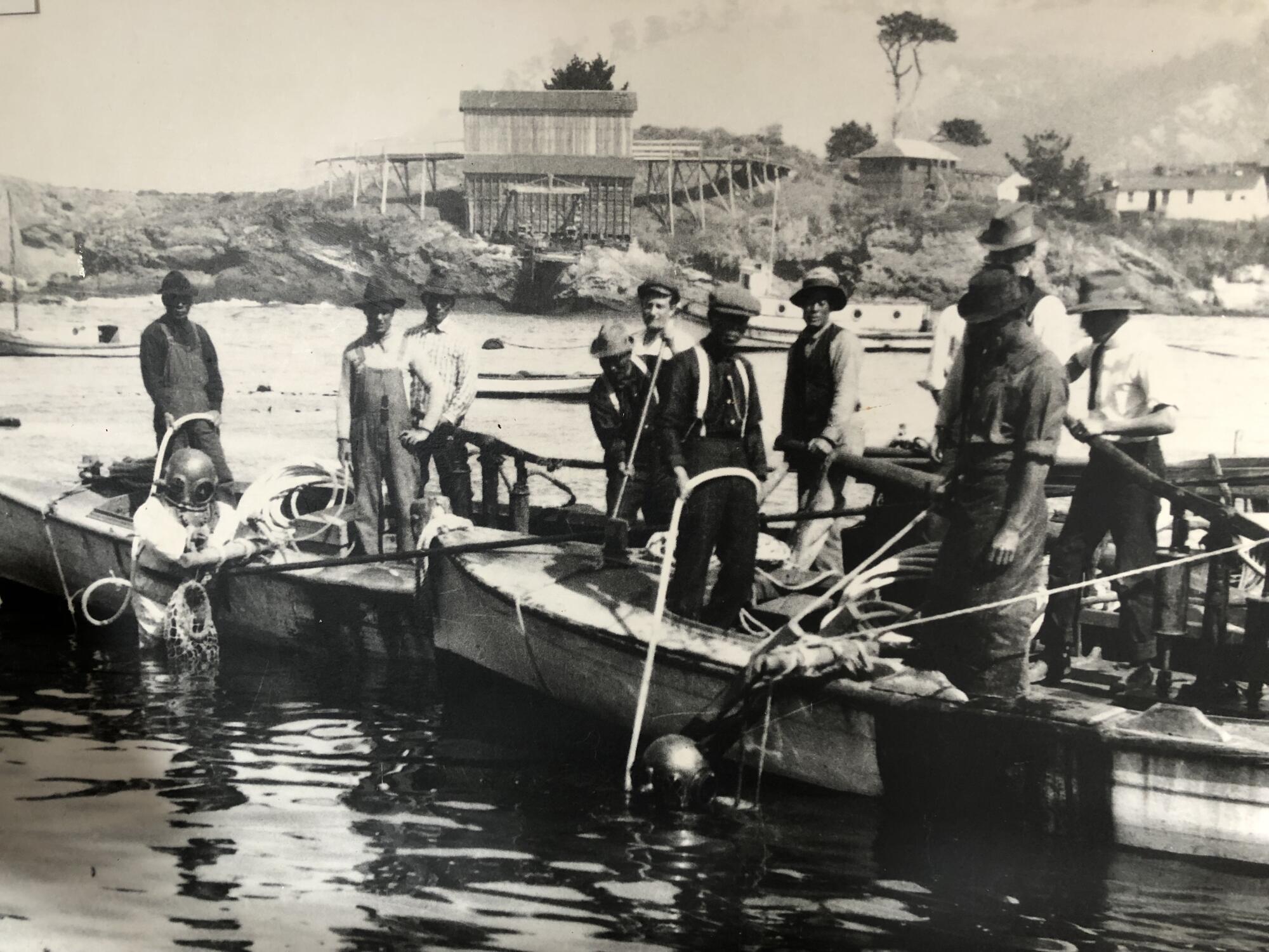 A black-and-white historic photo of several men standing on boats at water's edge as two divers emerge from the water.