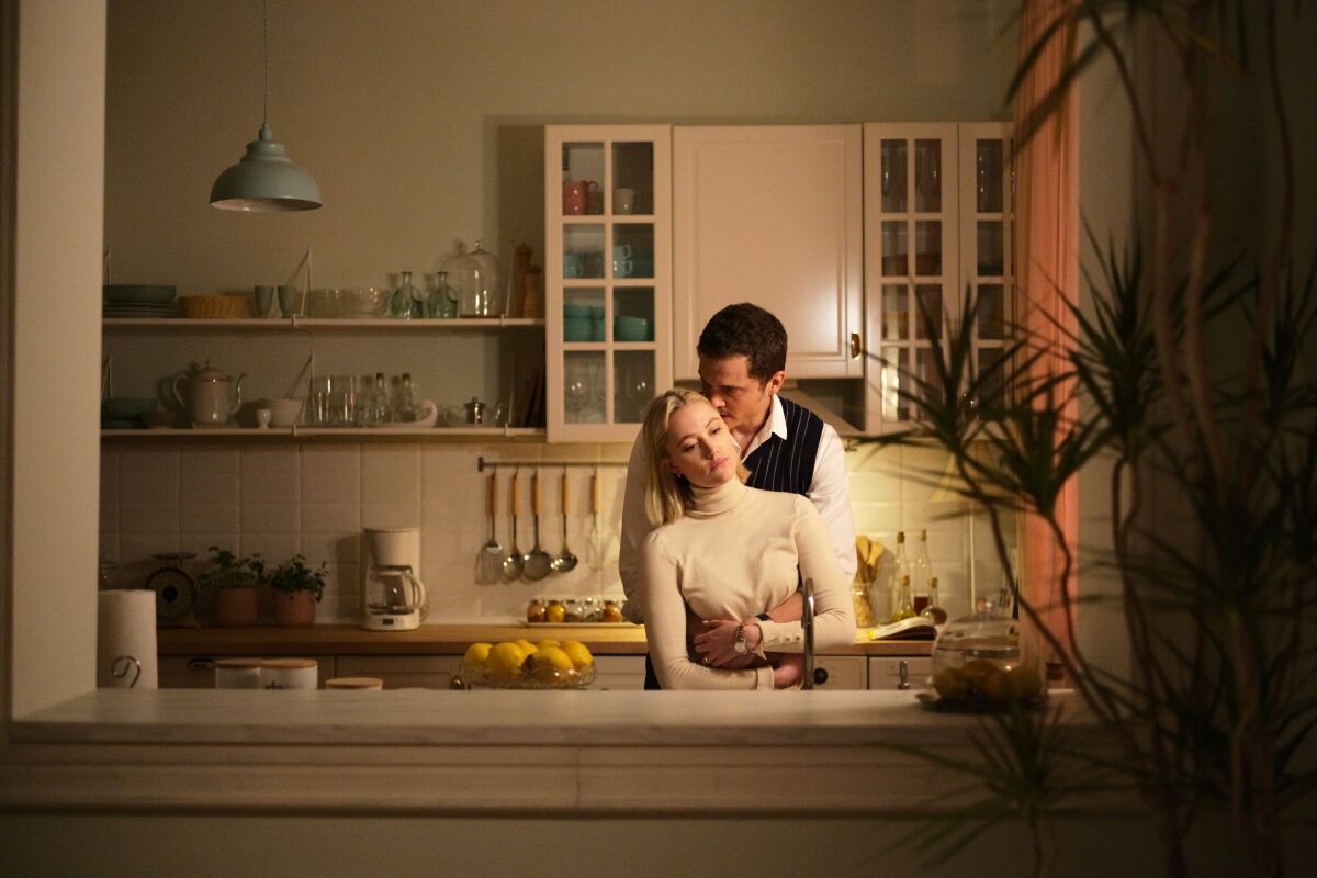 A man embraces a woman from behind in a kitchen in the movie “Watcher.”