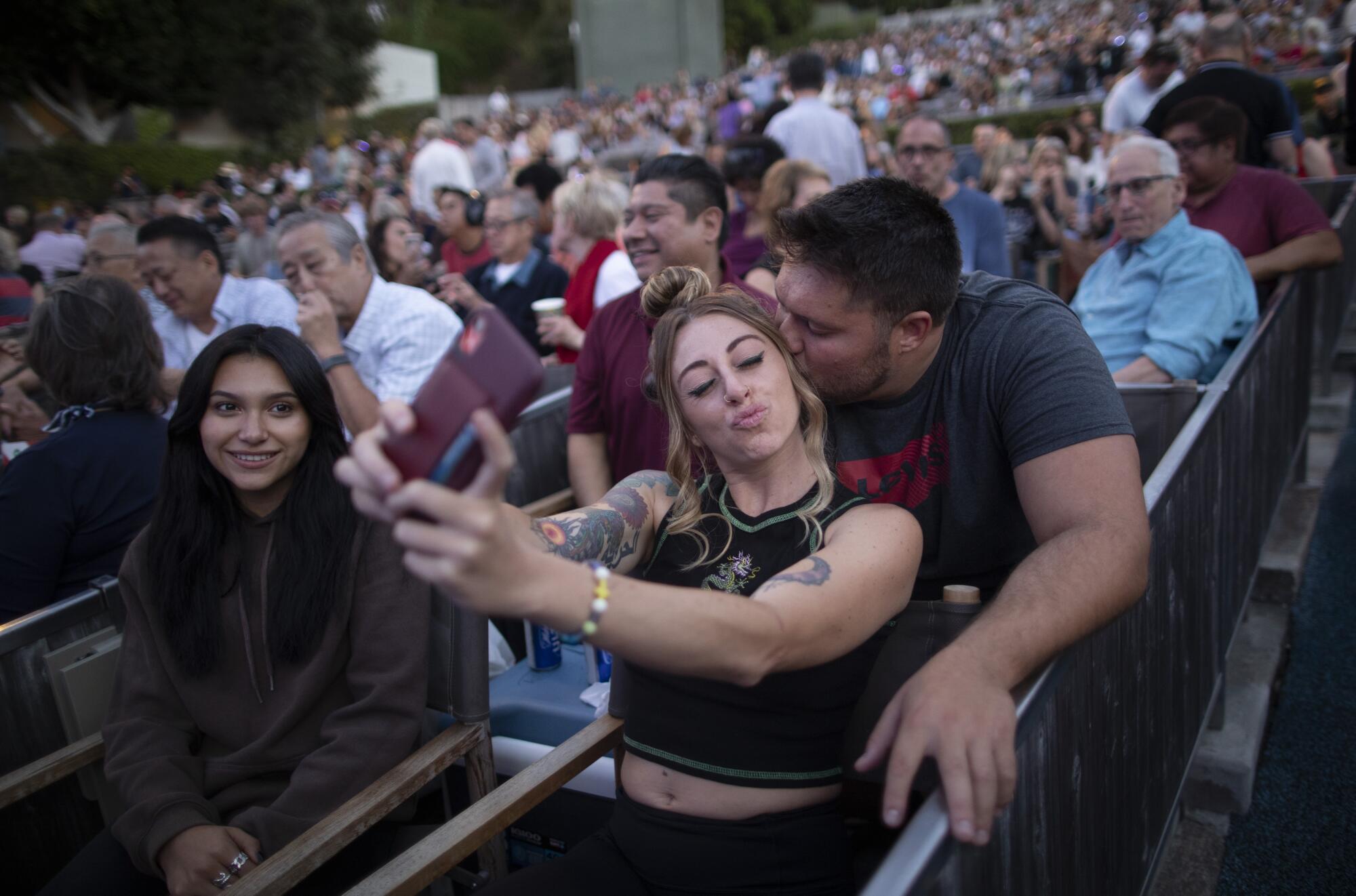 A man kisses the side of a woman's face as she takes their selfie.