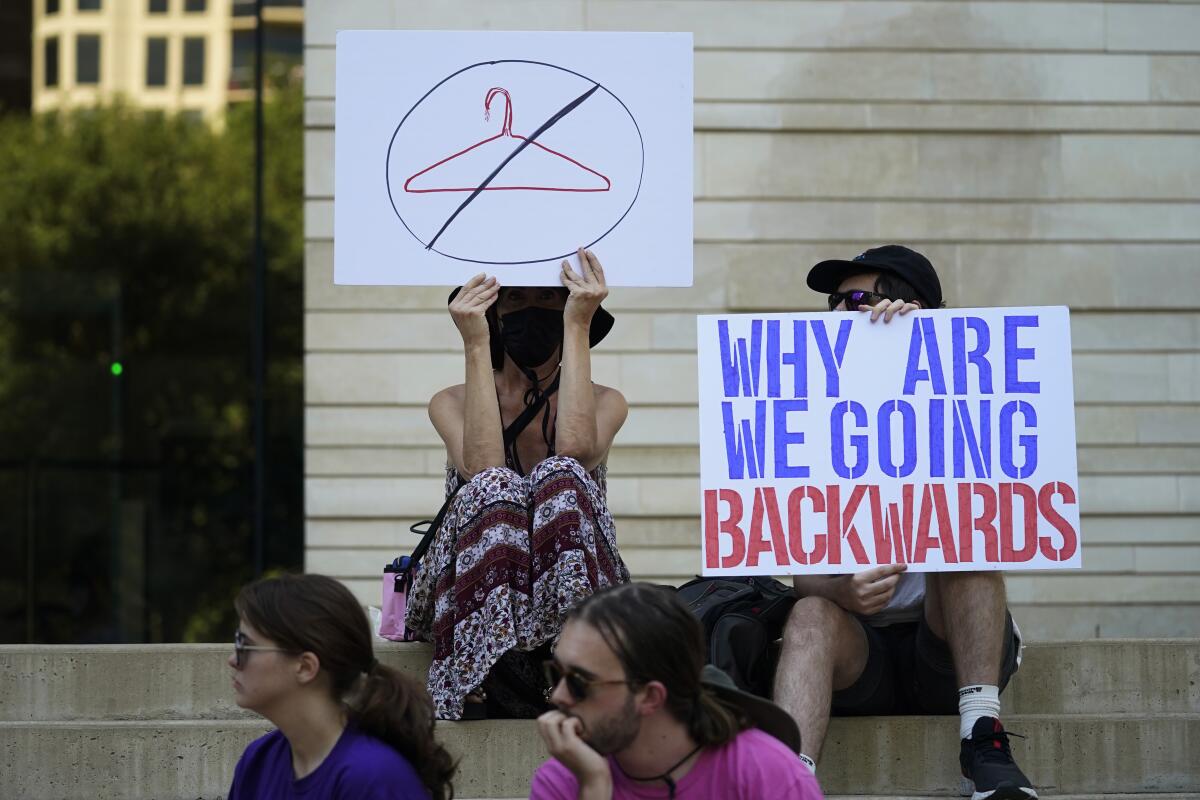 People hold signs on sitting on steps. One sign shows a hanger in a crossed-out circle.
