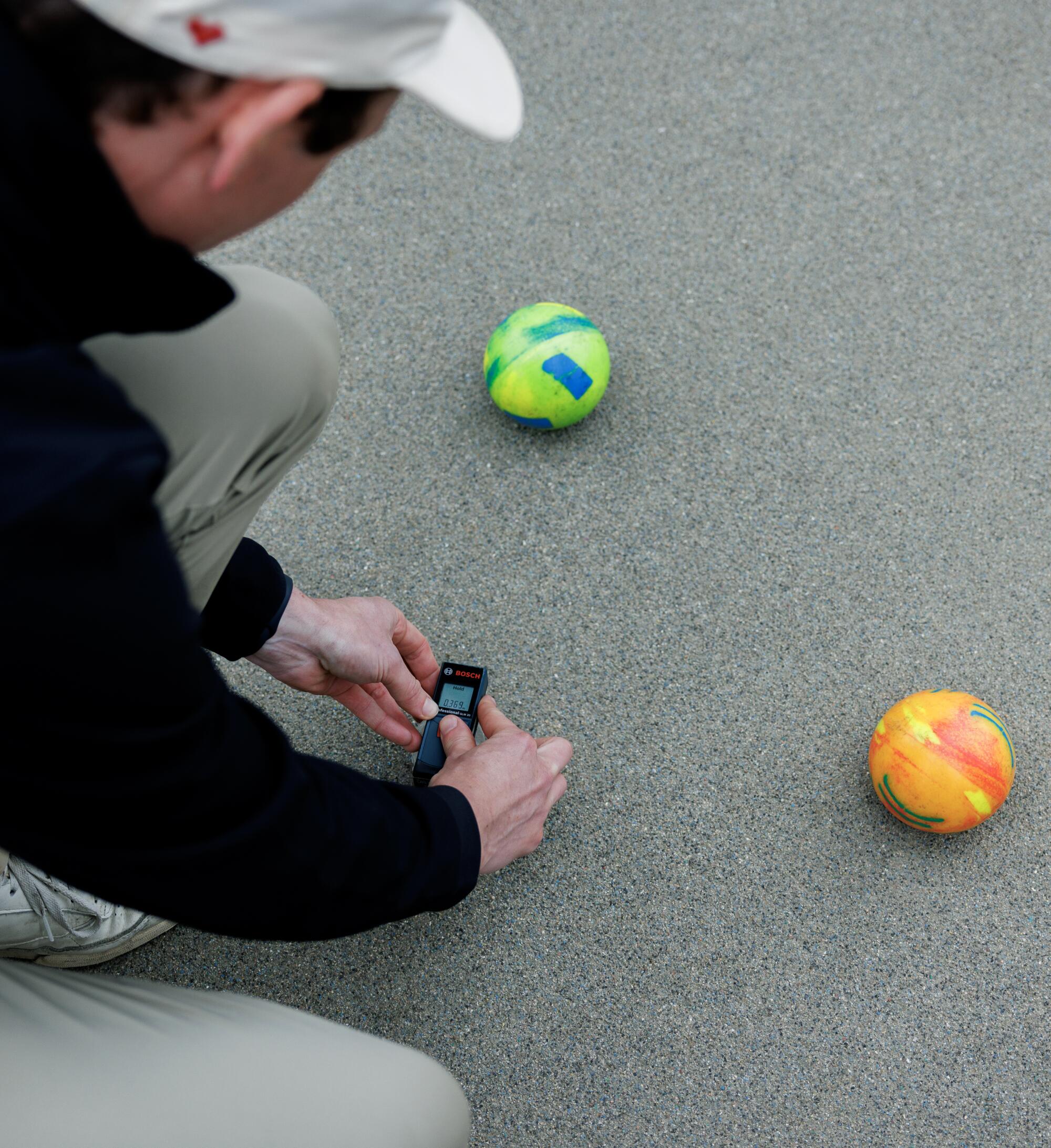 A man using a digital device to measure the distance between two bocce balls.