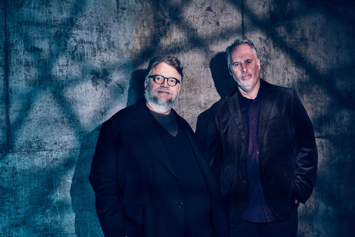 Guillermo del Toro and Mark Gustafson stand against a blue wall with shadows creating a grid of lines.