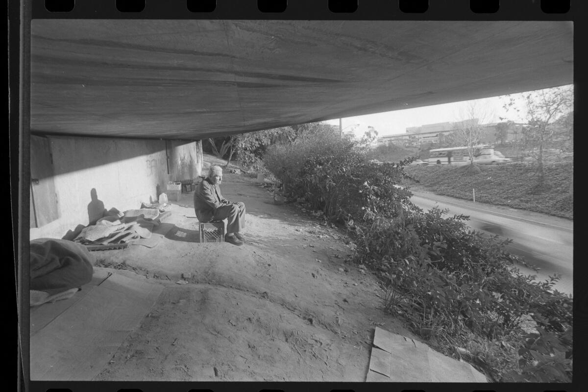 A man sits on a milk crate under an overpass watching the traffic on the Hollywood Freeway in 1985.