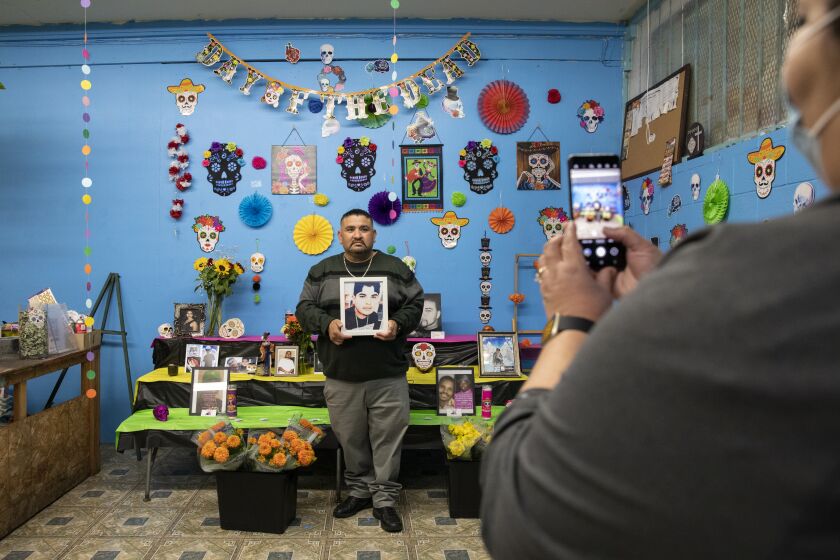 San Diego, CA - October 07: Carlos Munoz holds a picture of his late son as his wife, Elizabeth, takes a photograph during an event where families whose loved ones have been killed join The Union of Pan Asian Communities and Mums Flowers joined together to build an alter for Dia de los Muertos known as ofrendas to honor the dead at Mums Flowers on Thursday, Oct. 7, 2021 in San Diego, CA. This is the second year that the flower shop has had the alter and it will remain until November 3. The group has gotten to know each other and support each other through the years after the death of their children and other loved ones. "We create these healing spaces for out community," said Elizabeth Munoz. "We aren't grieving together, we're healing together." Their son Juan Carlos Munoz Jr. was shot and killed in 2015 in National City. (Ana Ramirez / The San Diego Union-Tribune)