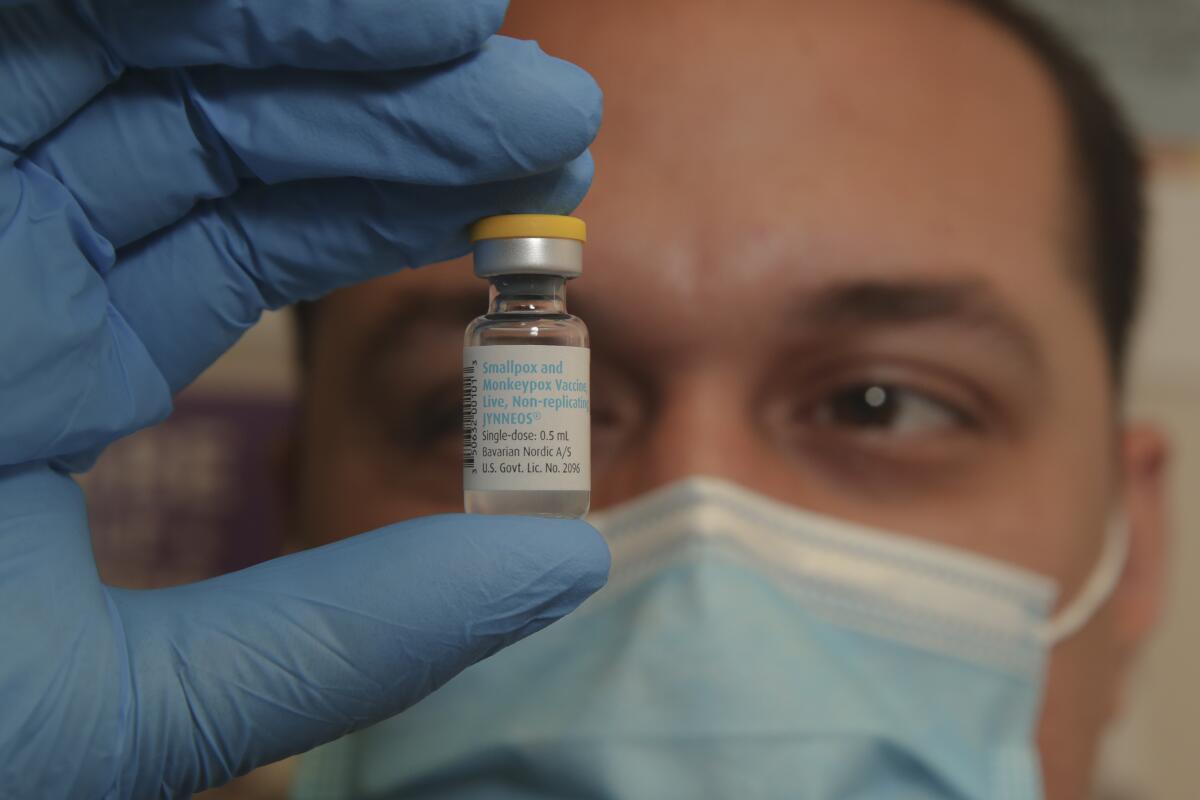 A gloved hand holding a vial in front of a face wearing a surgical mask.
