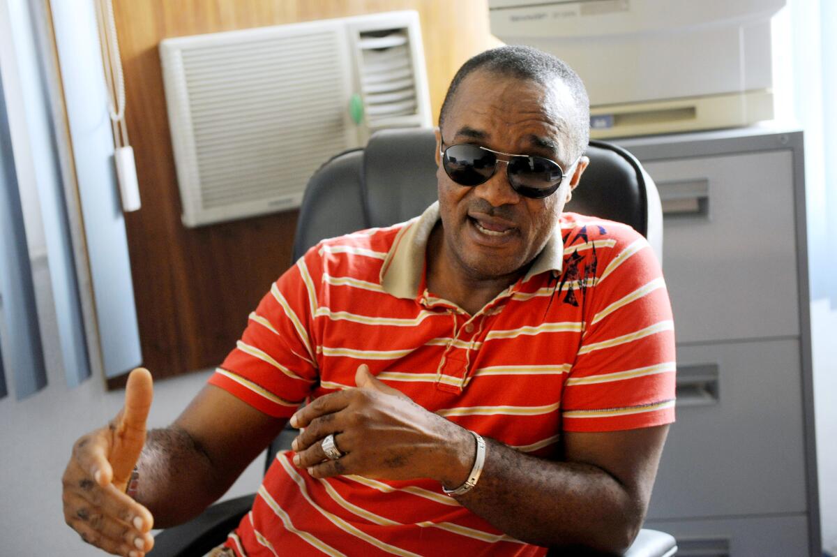 A man speaks and gestures while sitting in a chair, wearing sunglasses and a striped red shirt.