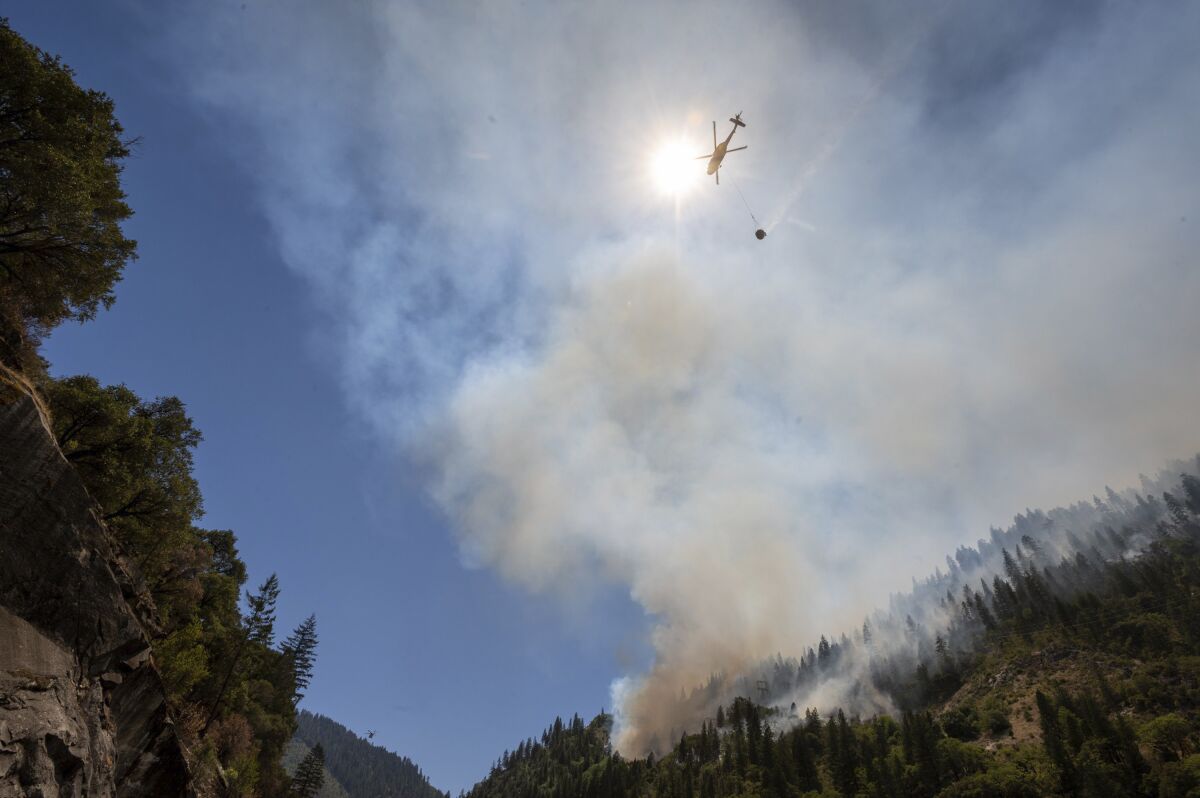 A bucket dangles from a helicopter approaching a wildfire