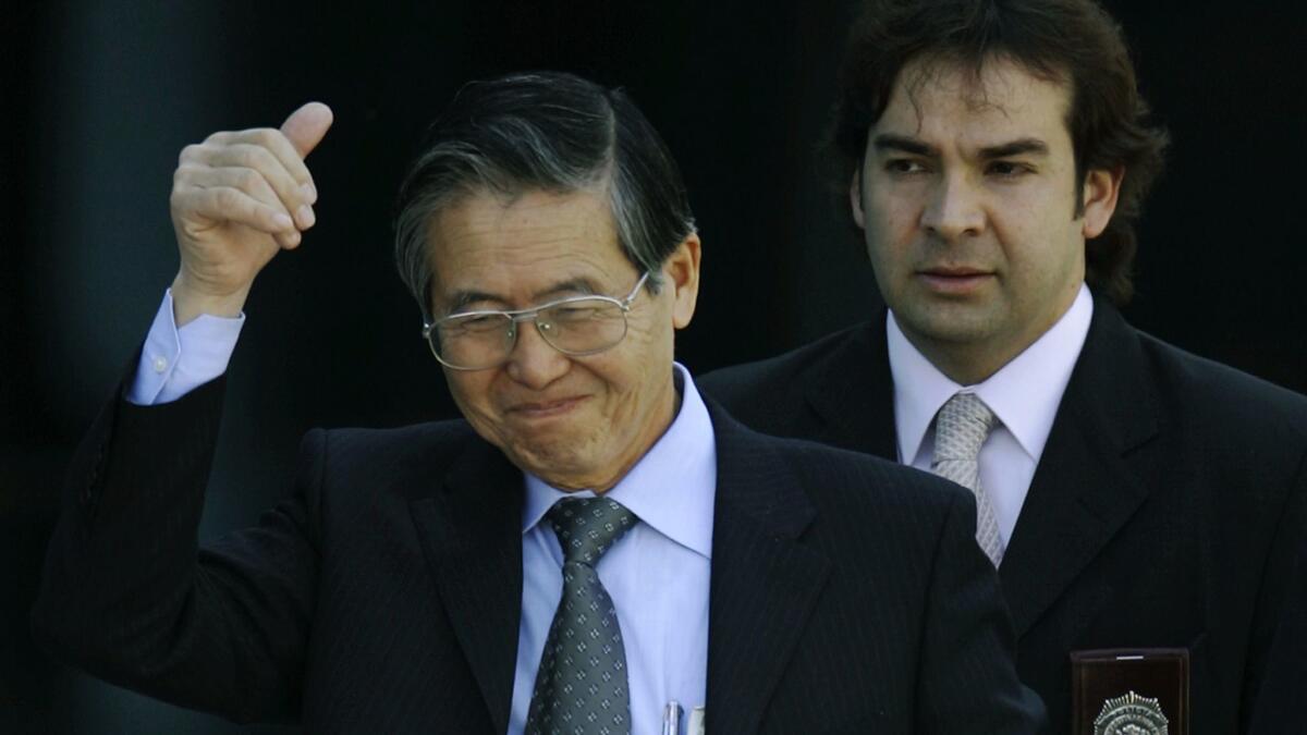 A man wearing glasses, in a dark suit and gray tie, smiles as he lifts a hand up