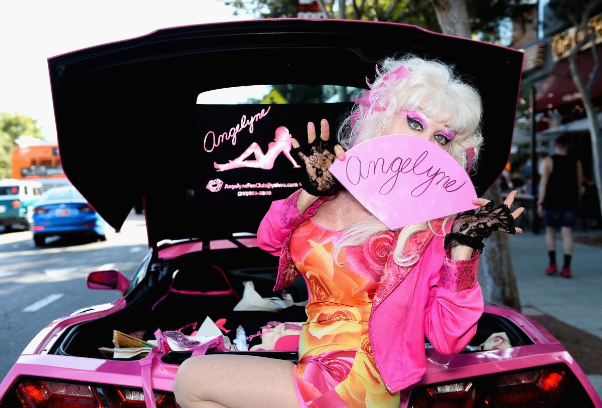 A very blond woman holding a pink fan in front of her face that says "Angelyne"