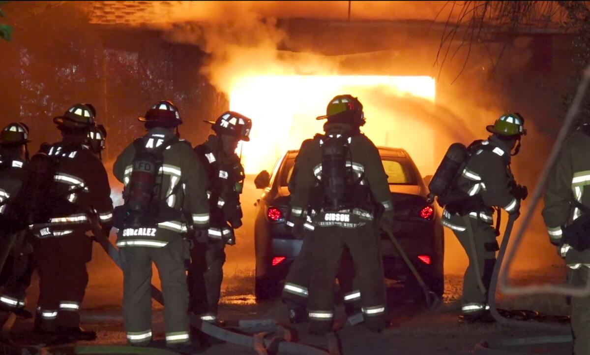 Firefighters in full gear stand outside a burning building before sunrise.