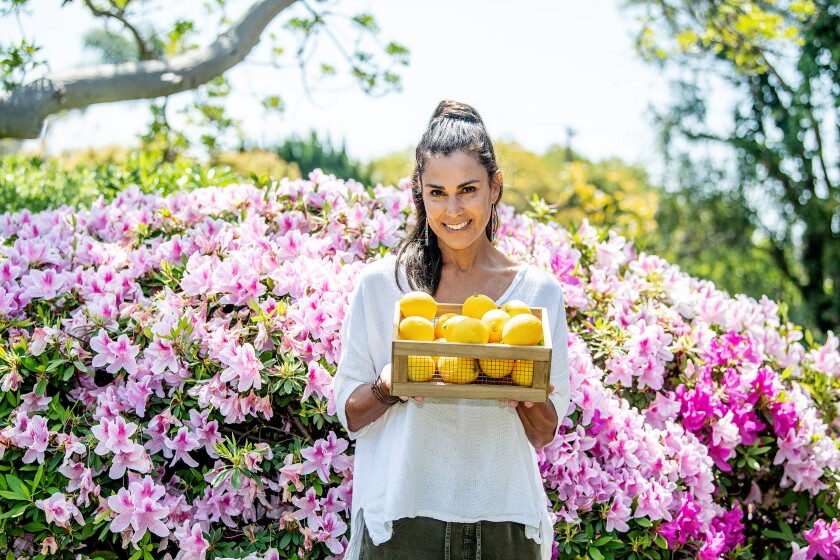 Alexandra Dorros holds a crate of lemons and stands in front of a flowering shrub