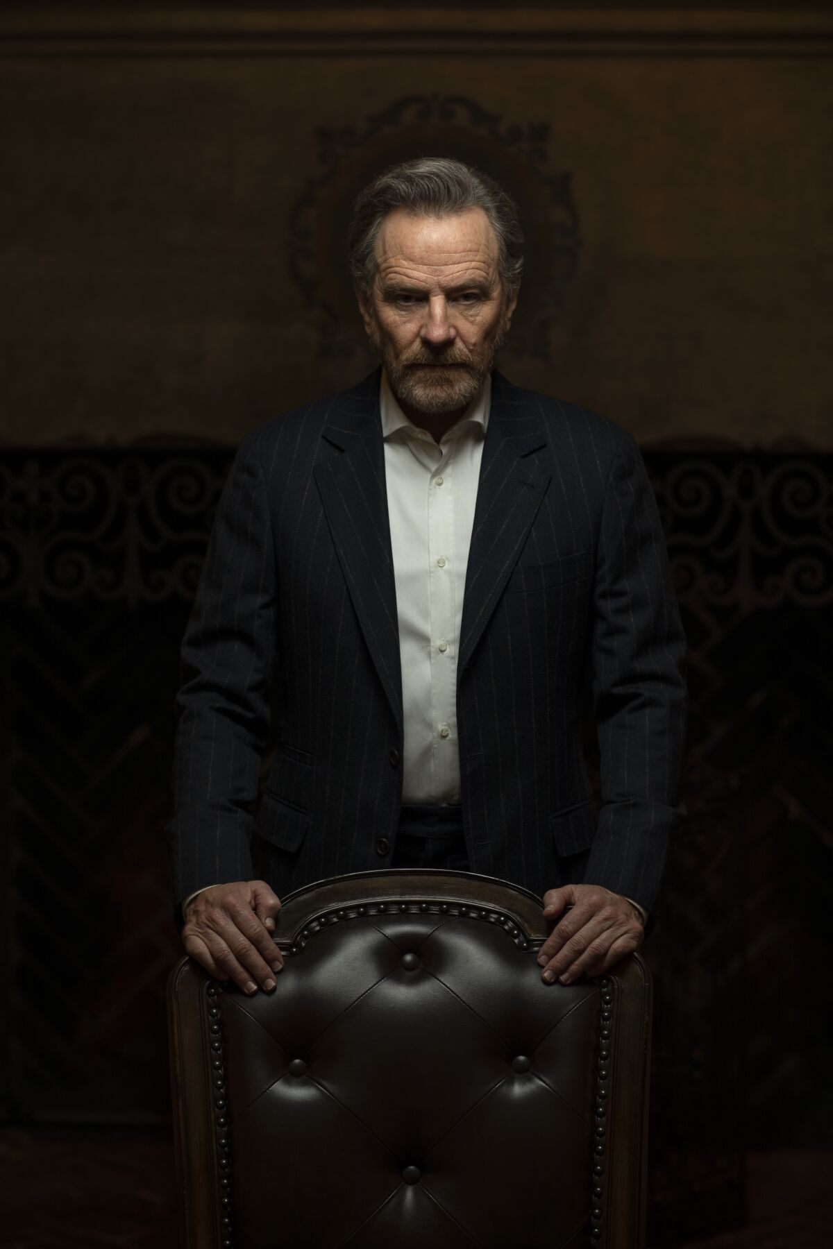 Actor Bryan Cranston, shrouded in shadows, stands dramatically behind a leather club chair.