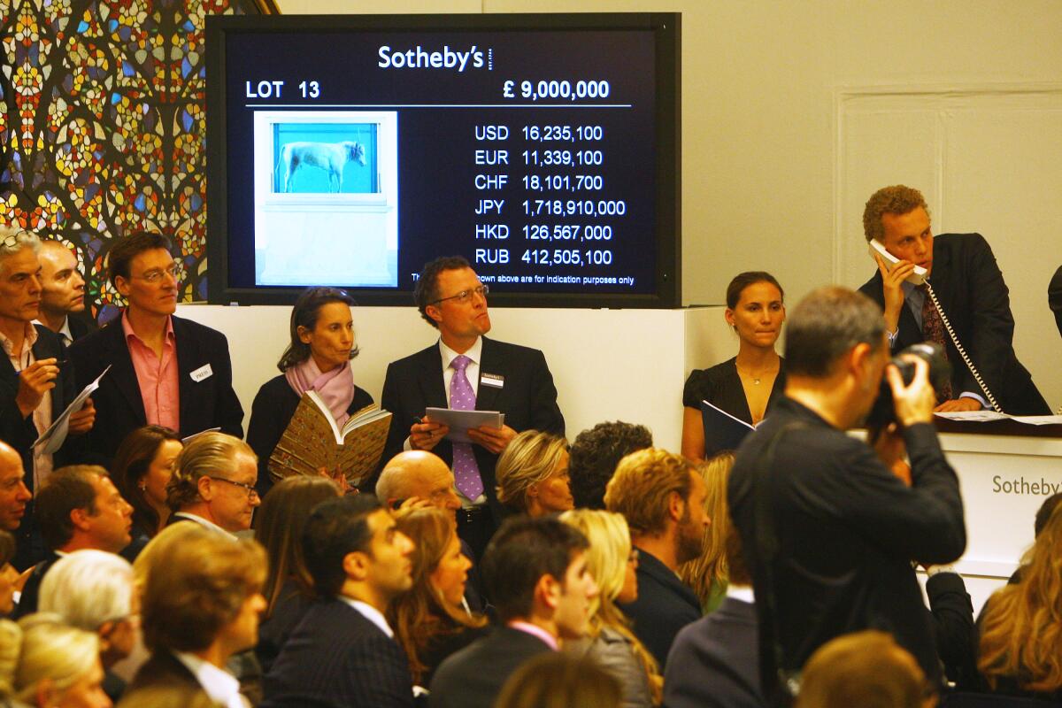 Auctioneers and patrons stand before a screen with a list of currencies in a crowded auction room 