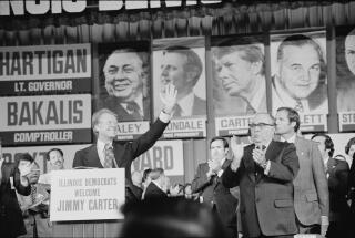 A man in a suit and tie smiles while waving as other men in suits and ties standing nearby applaud