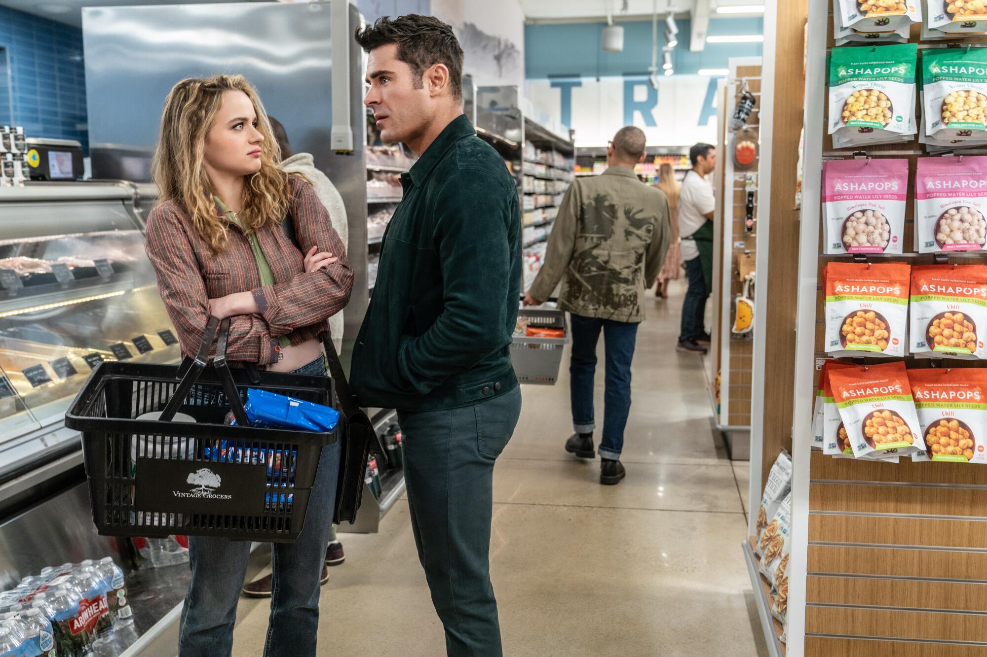 A woman stands near a grocery counter with a shopping basket, looking at a man.