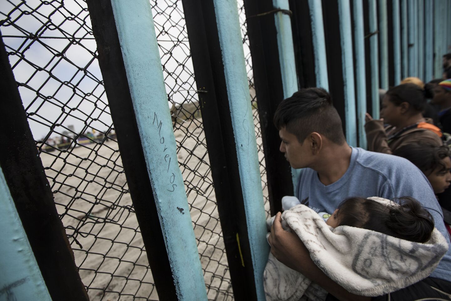 A member of the Central American migrant caravan, cradling a child, looks to the U.S. through the border wall at Tijuana.