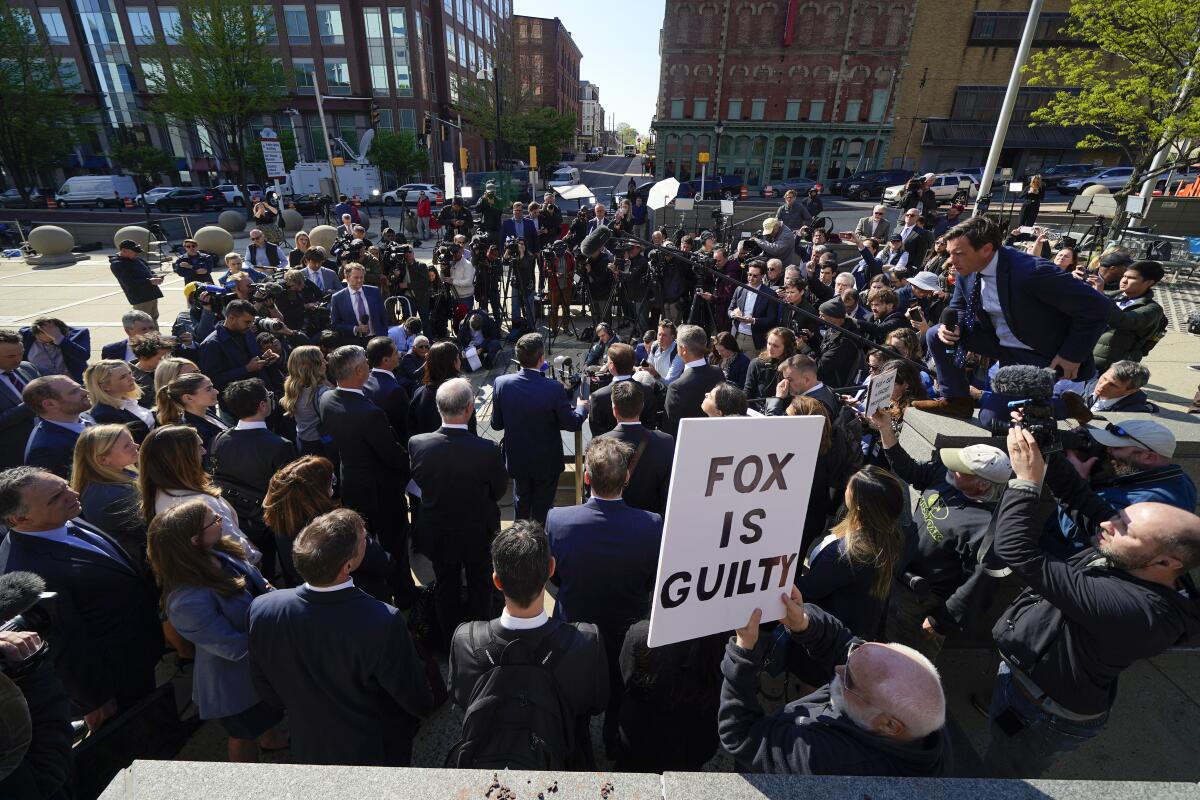 Attorneys speak to a crowd outside a courthouse, a member of which holds a sign reading "Fox is guilty."