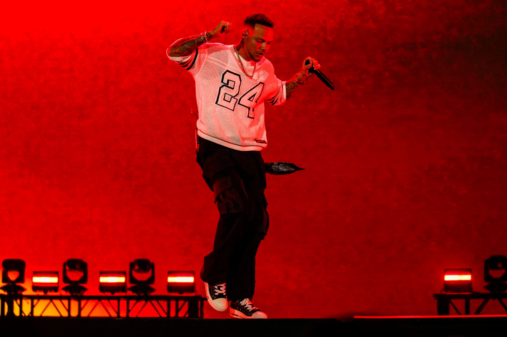 A man in a sports jersey and dark pants dances in front of a red background on stage.