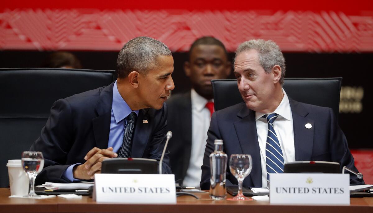 Two men confer while seated behind a table with microphones and place cards that read "United States"