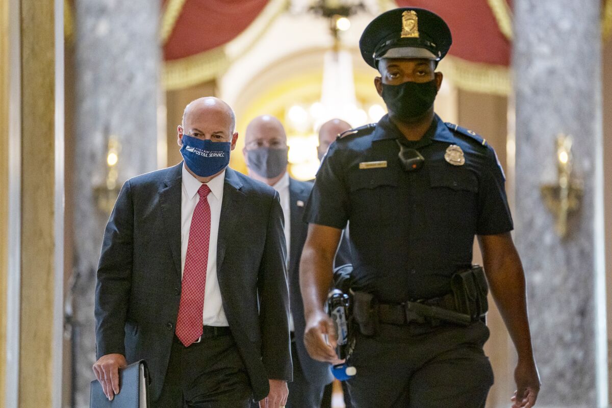 Postmaster General Louis DeJoy, wearing a suit and a mask, is escorted by a Capitol police officer