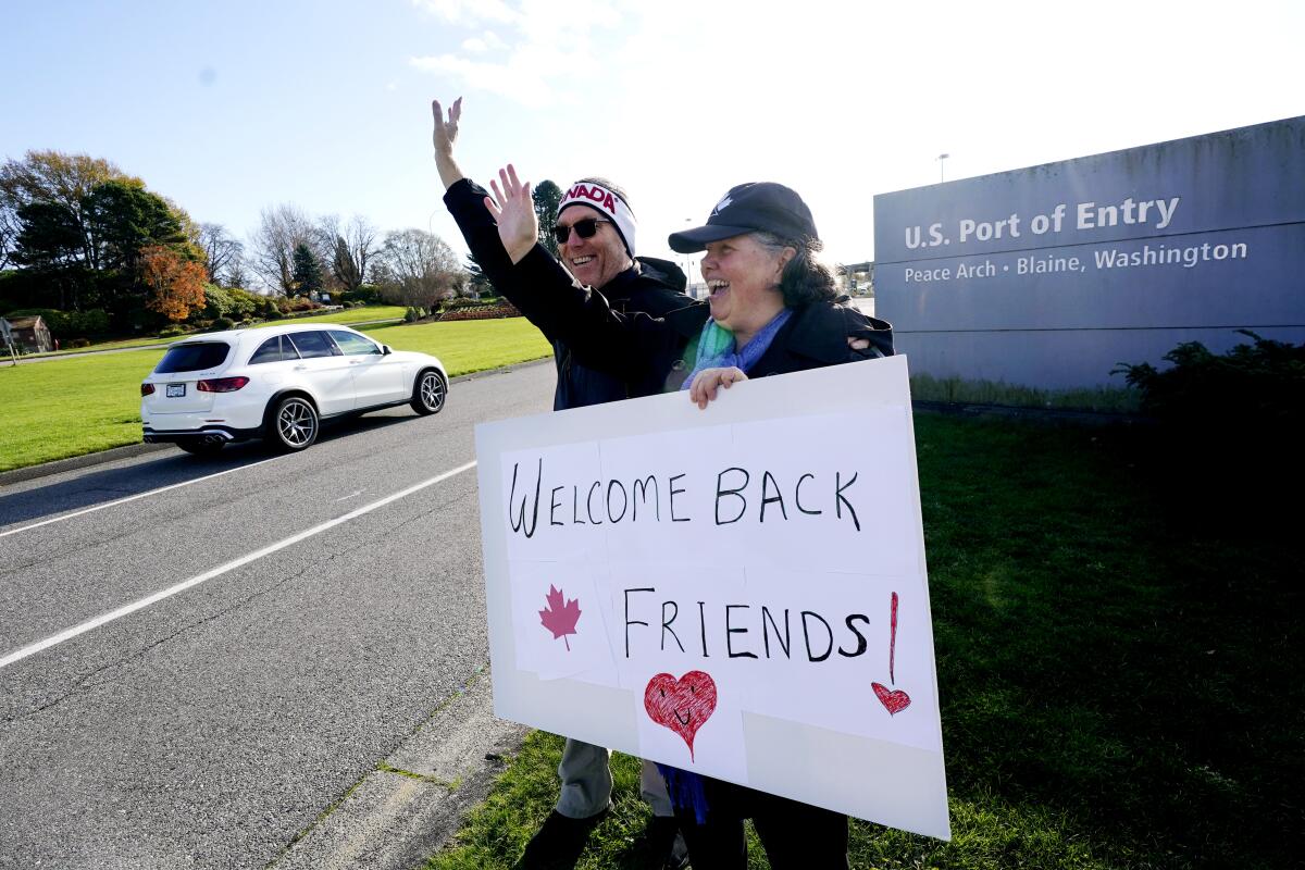 A man and woman stand in front of a U.S. port of entry sign holding a poster that says, "Welcome back friends!"