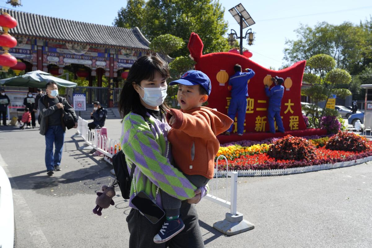 A woman wearing a mask carries a child past workers decorating a display for the upcoming 20th Party Congress in Beijing.