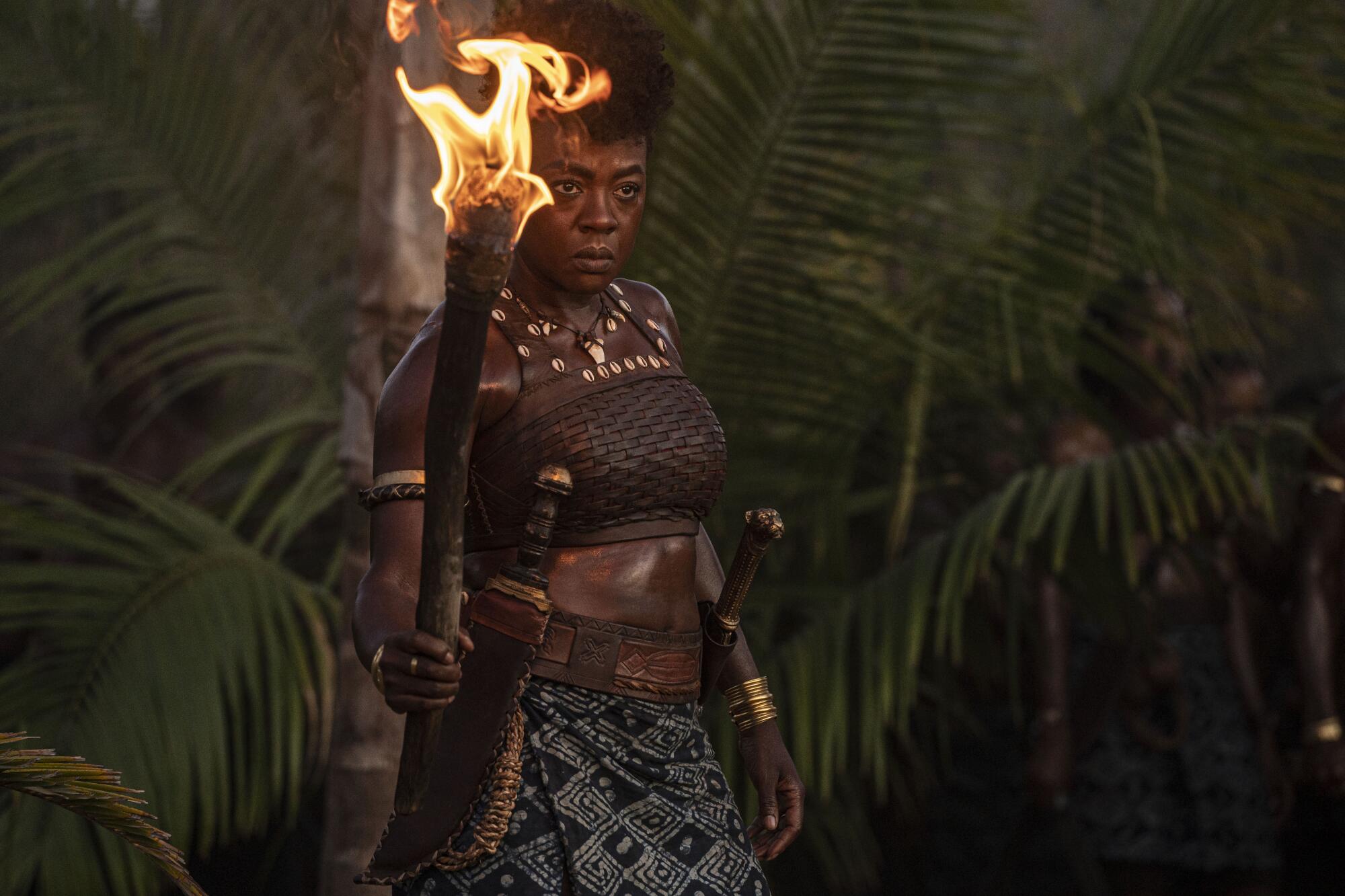 A woman warrior stands amid foliage holding a torch in a scene from "The Woman King."