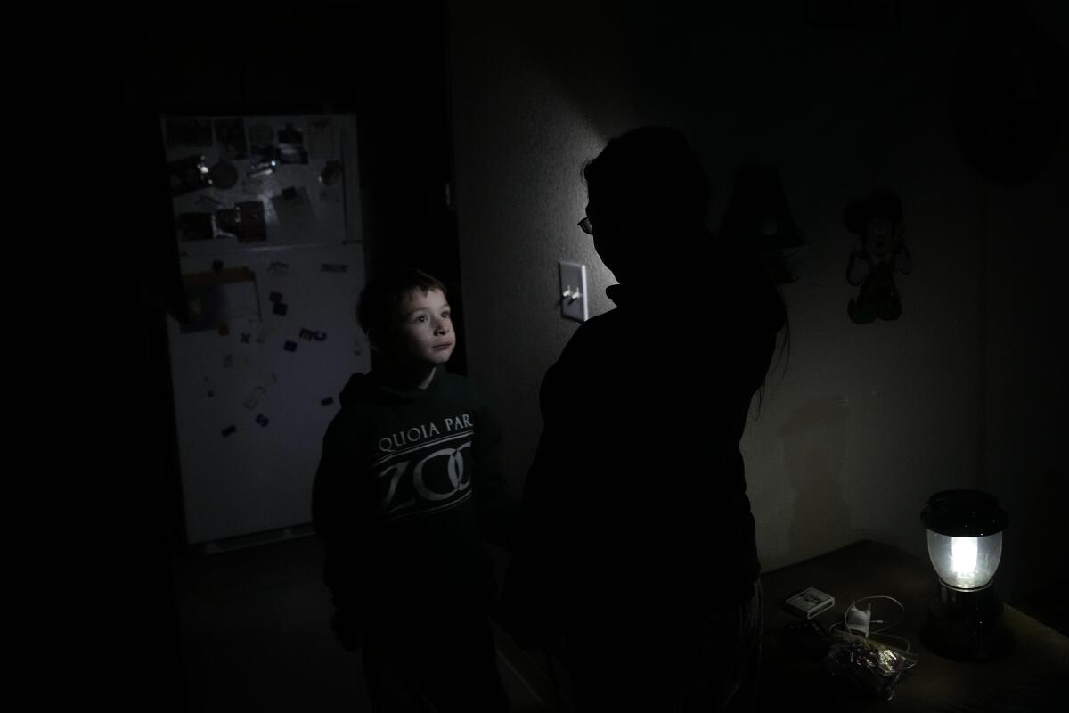 Standing in a dark kitchen, lit only by a small lamp, young boy looks up to a woman.