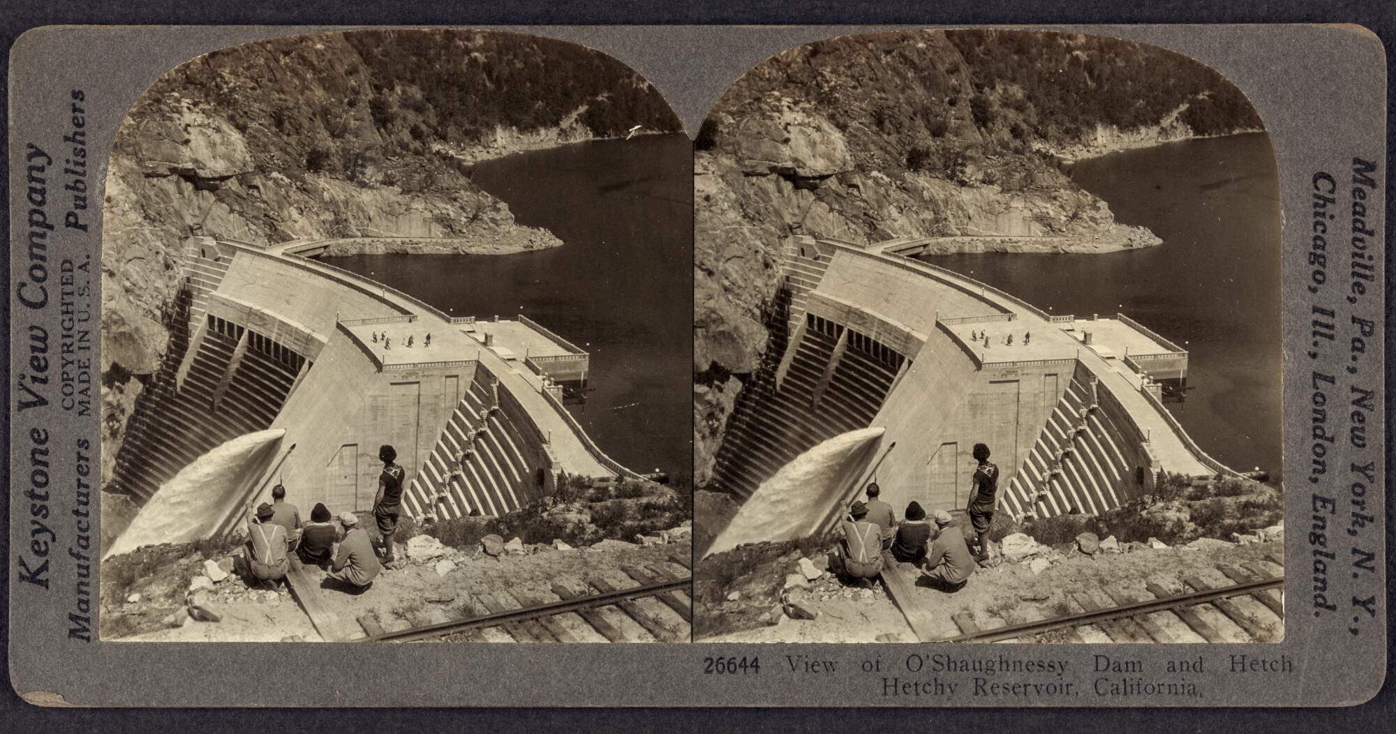 An old stereograph showing a small group of people next to railroad tracks atop a hillside overlooking the O'Shaughnessy Dam.