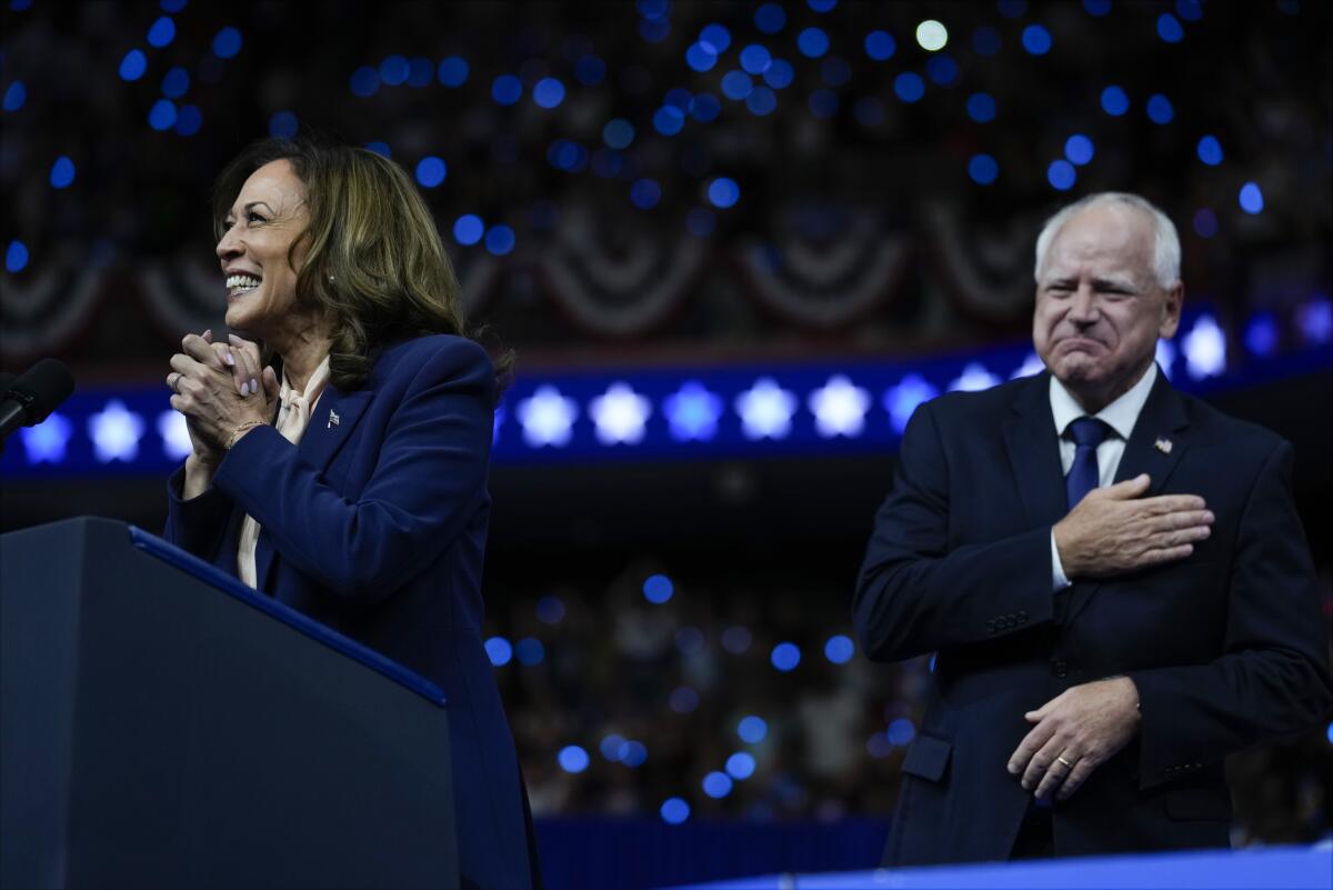 Kamala Harris at a lectern while Tim Walz holds his hand over his heart at an arena rally