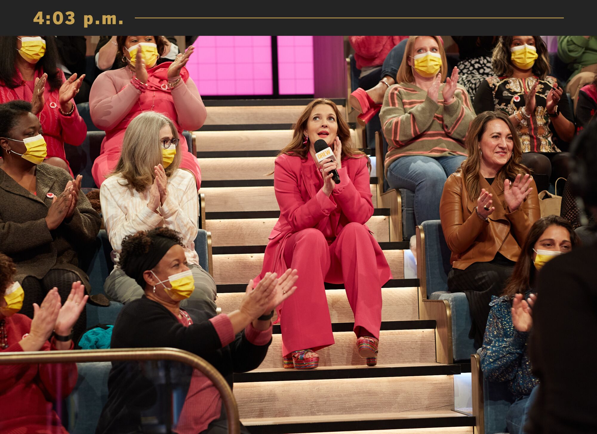 Drew Barrymore, wearing a pantsuit, sits on the steps of her talk show set amid the audience.